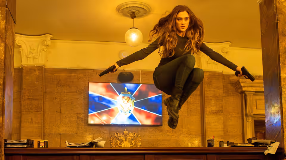 Renee (Lizzy Greene) leaps over a desk, holding pistols in an ornate room