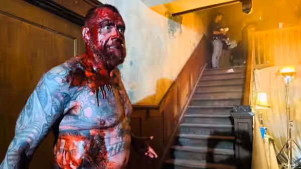 A bloodied man in a patterned shirt stands at the foot of a staircase as figures peer from above