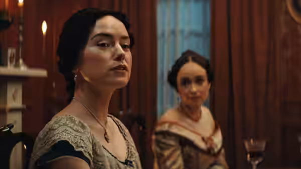 Two women in period dresses sit in a warmly furnished room