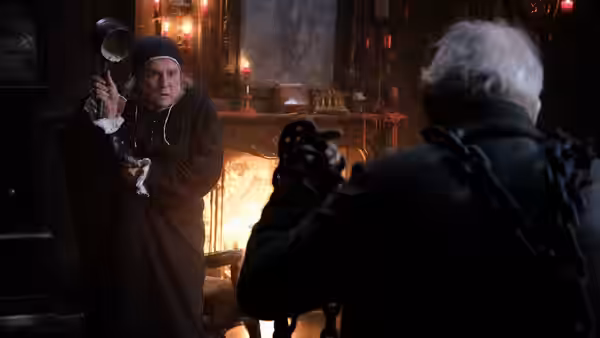 A wary man grips a candlestick beside the fire as a white-haired figure faces him