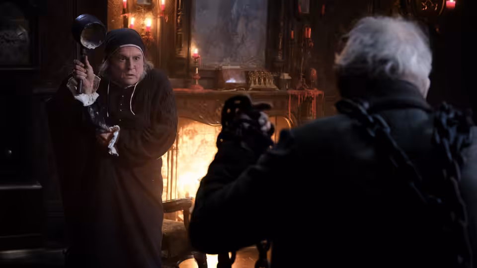A wary man grips a candlestick beside the fire as a white-haired figure faces him