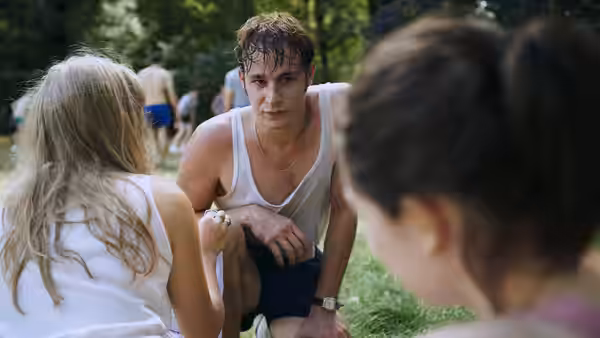 A young man crouches to speak with two women outdoors among trees