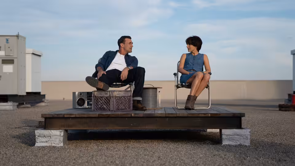 Lucy Cola (Natalie Portman) and Mark Goodwin (Jon Hamm) sit together on a rooftop platform