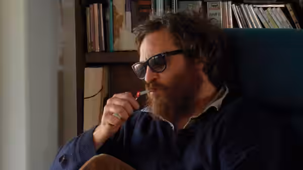 Joaquin Phoenix sits before bookshelves, holding a small object near his mouth