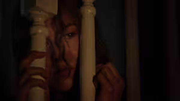 A woman peers anxiously through wooden bedposts in a dark room