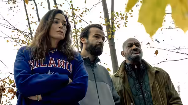 Three adults stand beneath autumn trees, looking down at something off-frame