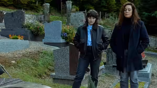 Two women stand among cemetery graves, looking toward something off-frame