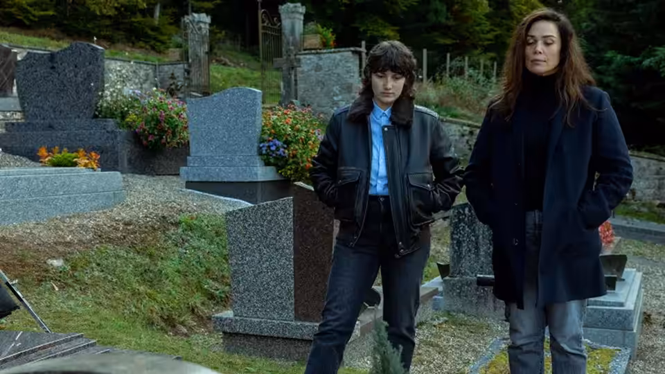 Two women stand among cemetery graves, looking toward something off-frame