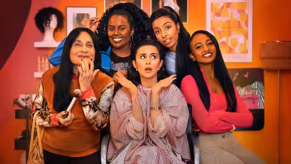 Five women gather around a seated woman, reacting with surprise in a colorful room