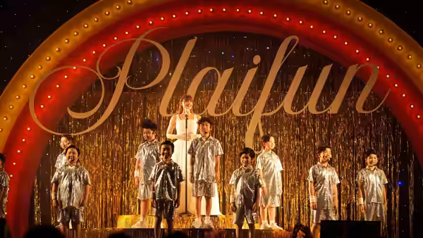 A group of performers sing onstage beneath a Plaifun sign and golden curtains