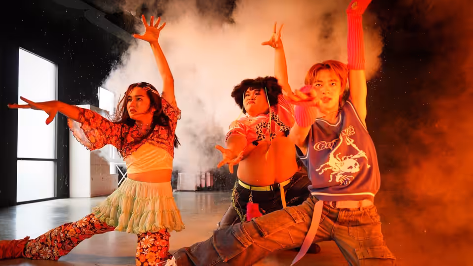 Three performers dance through smoke in colorful outfits inside a rehearsal space