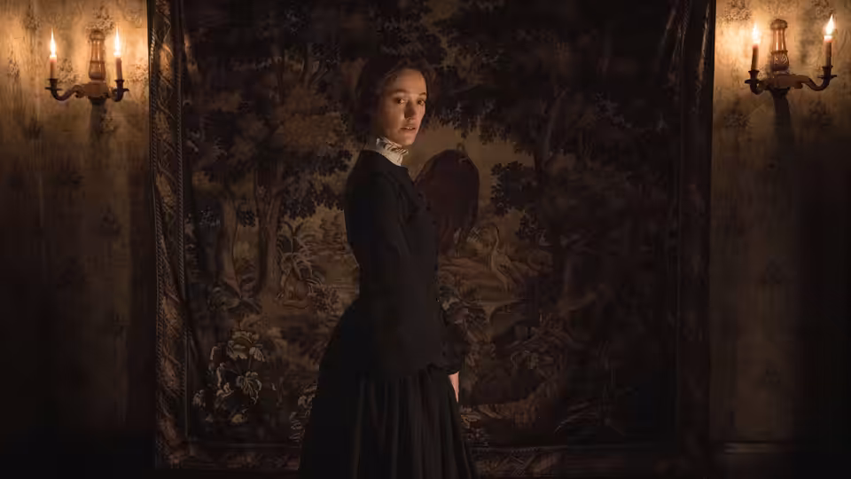 A woman in a dark Victorian dress turns toward the viewer in a tapestry-lined room