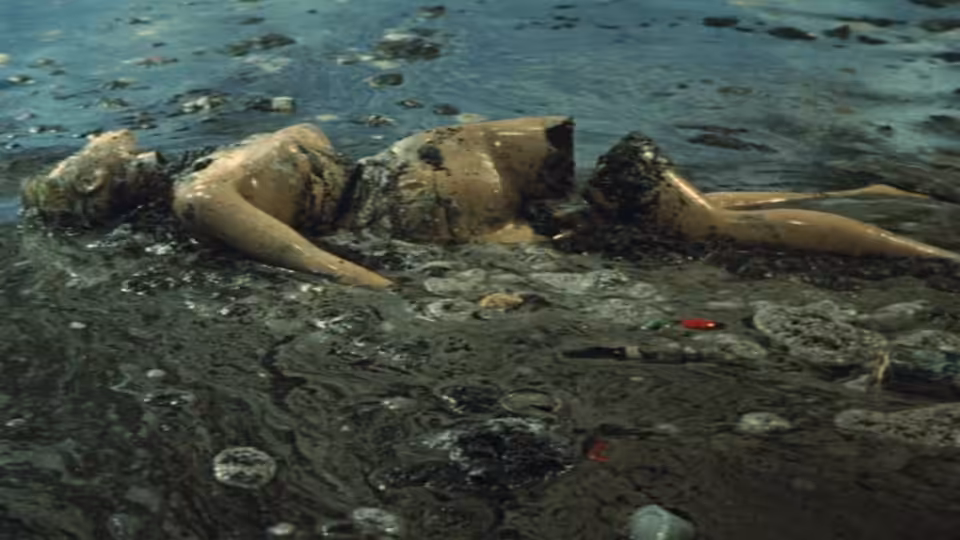 A person lies face down in polluted water strewn with debris