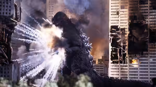 Godzilla towers between damaged city buildings as explosions erupt around him