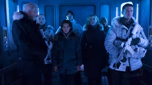 A group of bundled-up people stand inside a blue-lit aircraft cabin