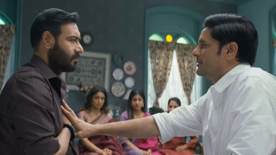 Vijay Salgaonkar (Ajay Devgn) faces a man pressing a hand to his chest as women watch
