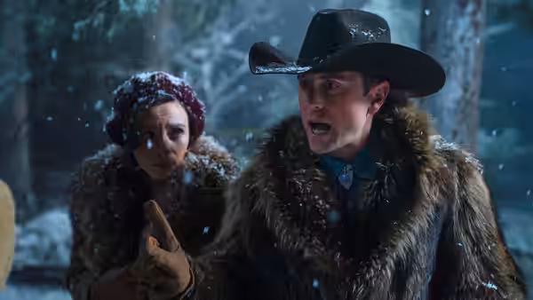 A fur-clad man gestures beside a bundled woman in a snowy forest