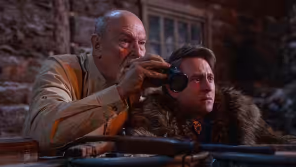 An older man peers through a viewing device beside a fur-clad younger man