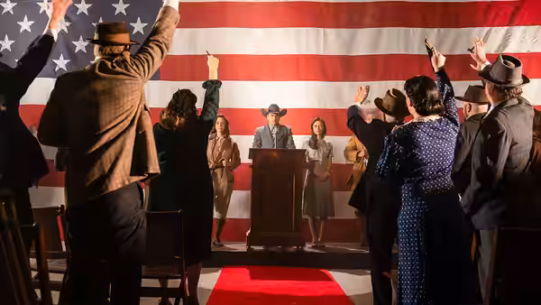 A speaker addresses a crowd raising their hands beneath a large American flag
