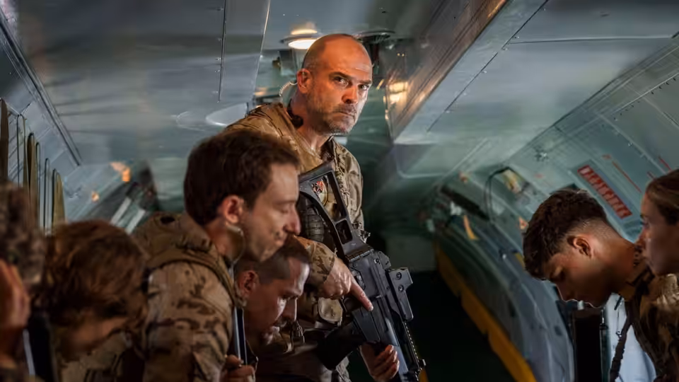 Armed men crouch inside an aircraft cabin as a bald soldier holds a rifle