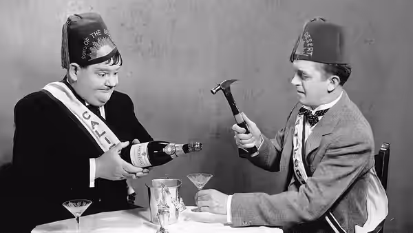 Stan (Stan Laurel) and Ollie (Oliver Hardy) sit at a table in Sons of the Desert caps and sashes