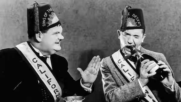 Stan (Stan Laurel) watches as Ollie (Oliver Hardy) drinks from a bottle at the table