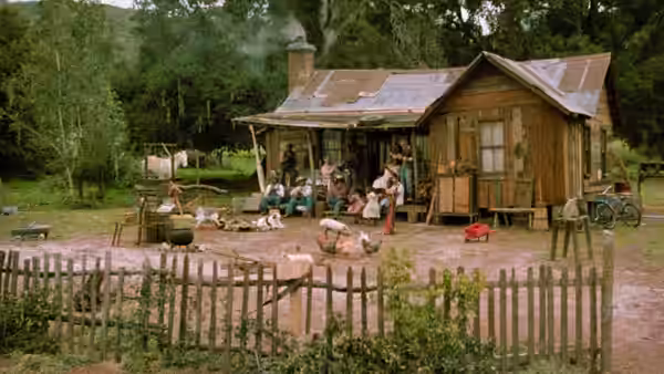 A family gathers outside a wooden cabin in a rural yard