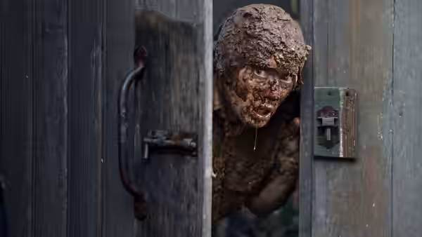 A mud-covered man peers through a partly opened wooden door