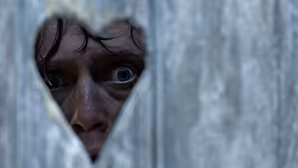 A frightened man peers through a narrow gap in a wooden wall