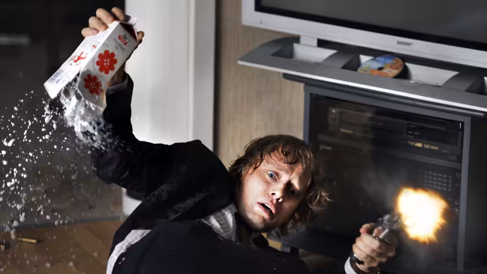 Roger Brown (Aksel Hennie) fires a gun while falling beside a kitchen oven
