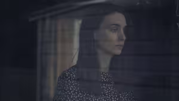 M (Rooney Mara) looking out a window from inside a dark room