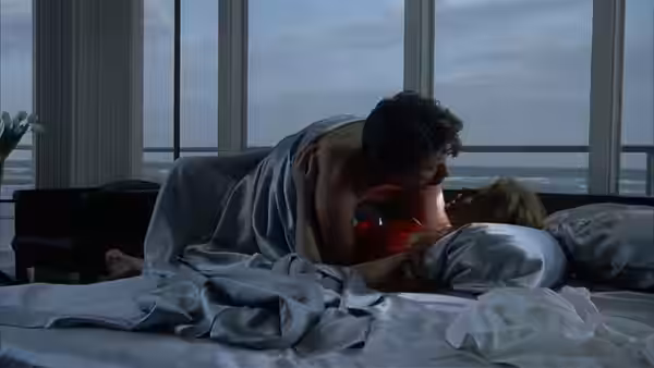 Martin leans over Laura in bed beside floor-to-ceiling windows overlooking the water