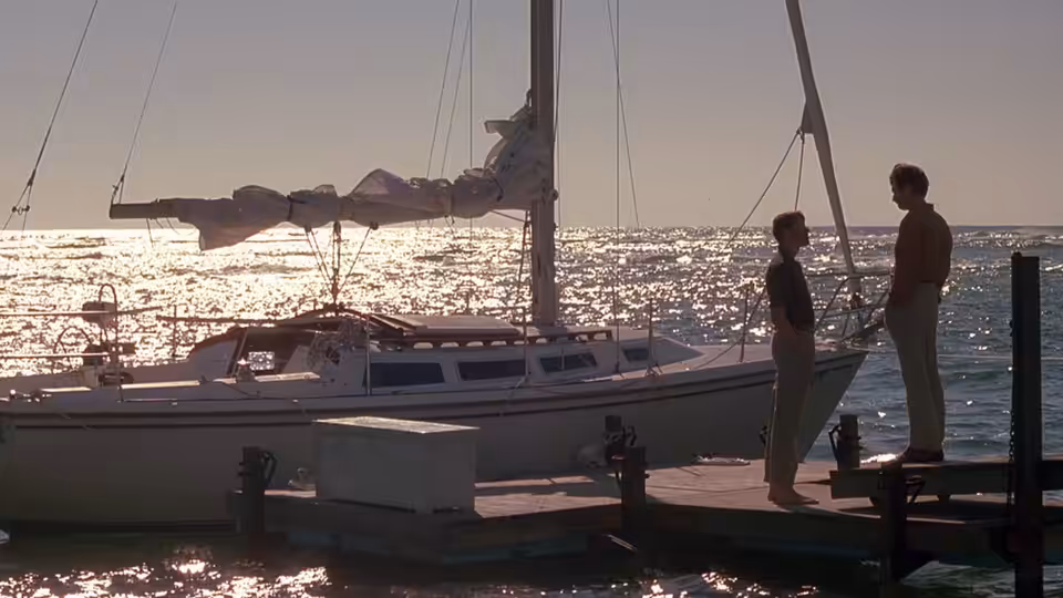 A couple stand together on a marina dock beside a sailboat at sunset