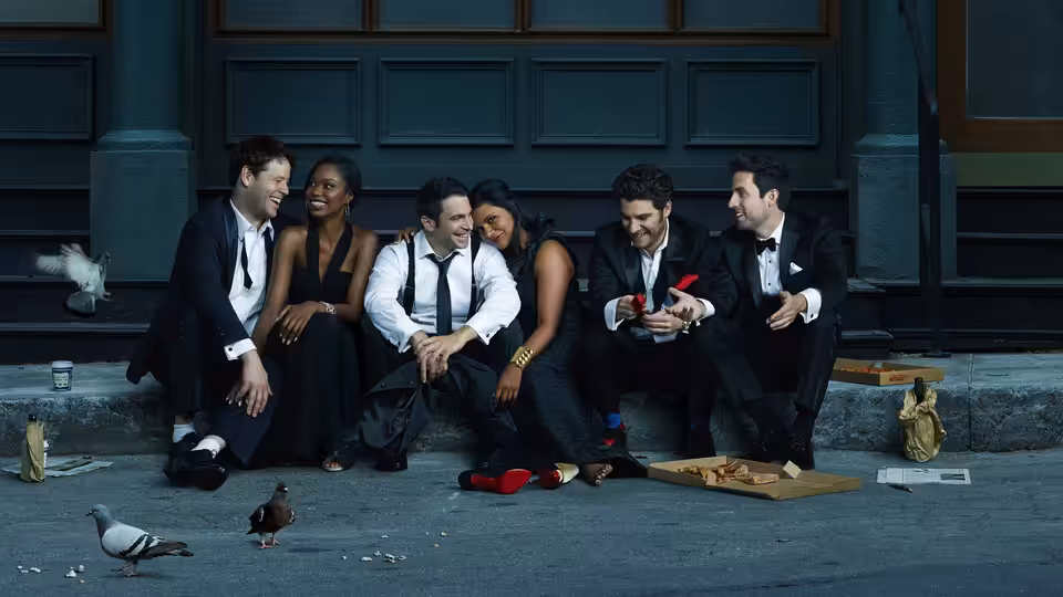 Six formally dressed people sit together on a city sidewalk as pigeons gather nearby