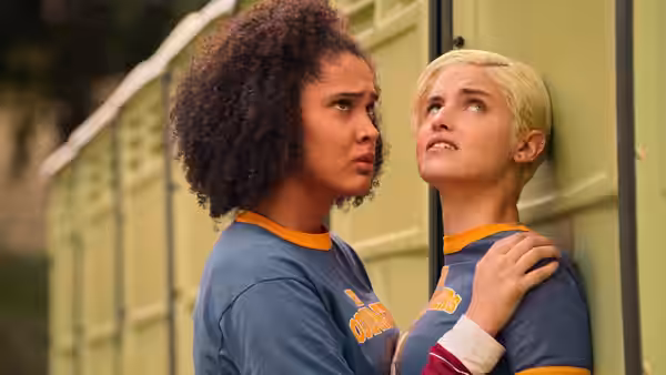 A curly-haired student comforts a blonde teammate beside a row of lockers