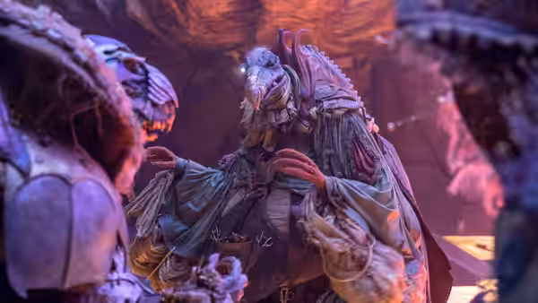 A robed, many-eyed creature gestures toward another figure in a glowing cavern