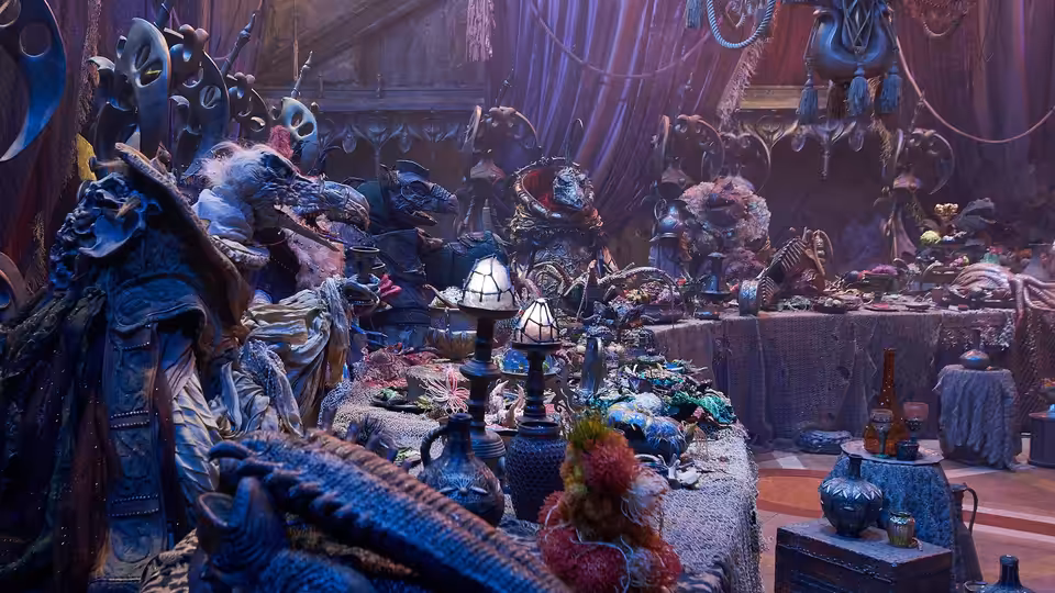 A crowded chamber of robed creatures gathered around a long table laden with food and vessels