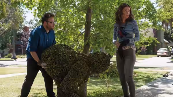 Mac trims a hedge while Kelly stands beside him on the sidewalk