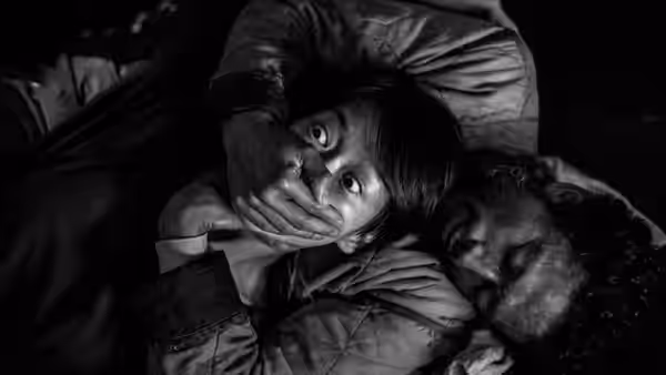 A man covers a woman’s mouth as they lie together on the ground