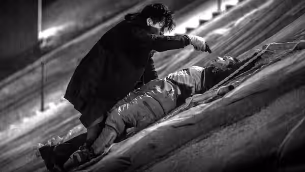A man kneels over another man on a sloping rooftop, holding a knife