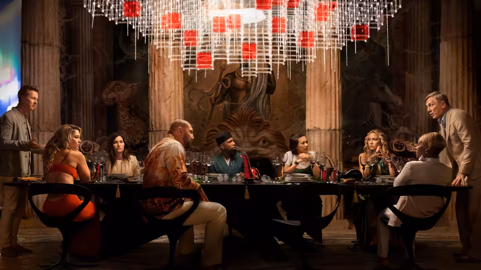 Benoit Blanc (Daniel Craig) and the guests gather around a long table beneath a chandelier