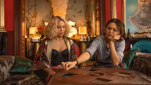 Birdie Jay (Kate Hudson) watches Peg (Jessica Henwick) reach across a wooden game table