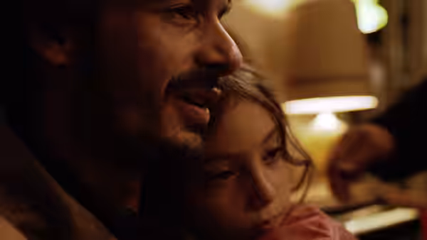 A bearded man holds Sol (Naíma Sentíes) close in a warmly lit room