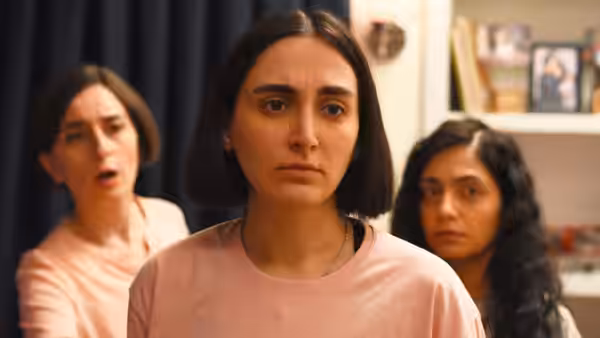 A young woman stands tensely between two women in a warmly lit room