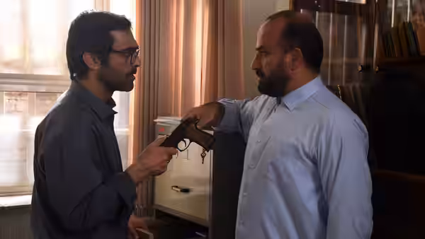 A bearded man offers a handgun to a bespectacled man beside an office cabinet