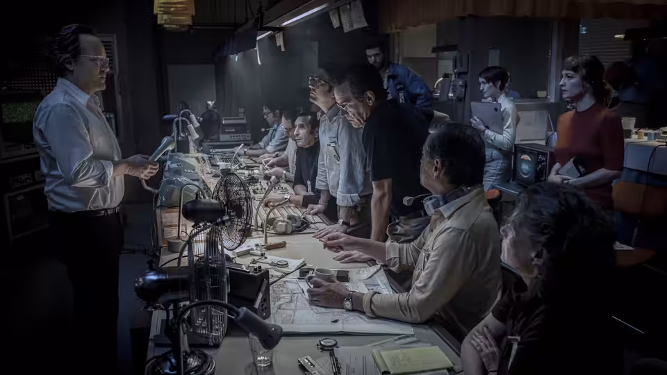 A newsroom team gathers around a crowded desk as a man gestures to the staff