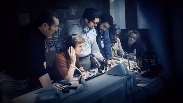 A woman wearing headphones takes a call while colleagues crowd around the control room desk