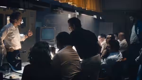 A man addresses seated colleagues beside a bank of monitors in the control room