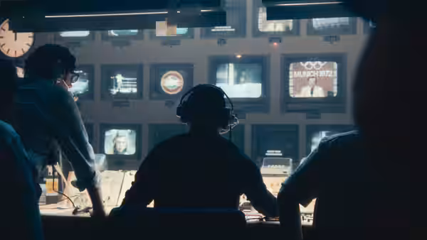 Three operators sit at the control desk watching broadcasts on a wall of monitors