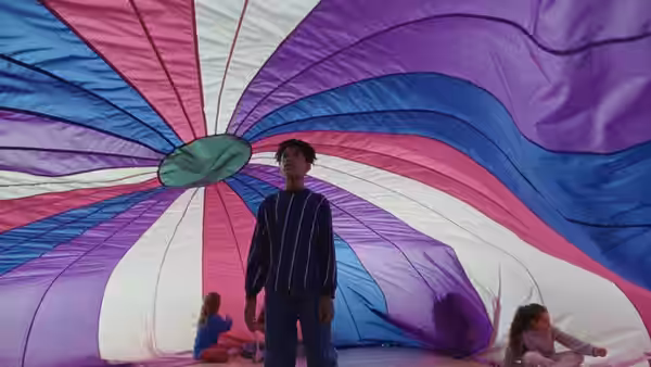 A young person stands beneath a vast, colorful parachute as others sit nearby